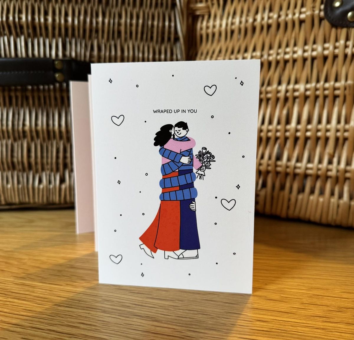 Greeting card with illustration of two people hugging, surrounded by hearts, on a wooden surface.