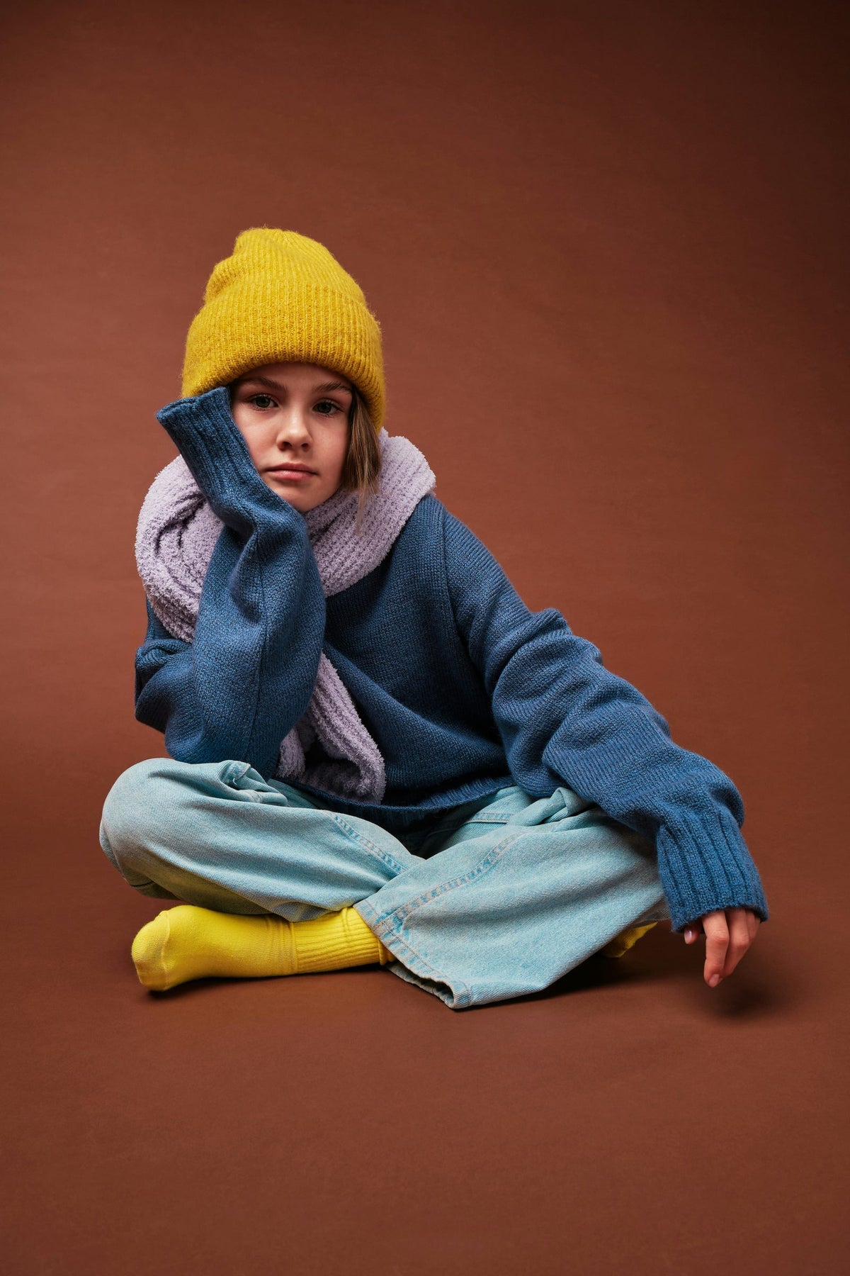 Child in a yellow beanie and blue knit jumper sitting against a brown backdrop