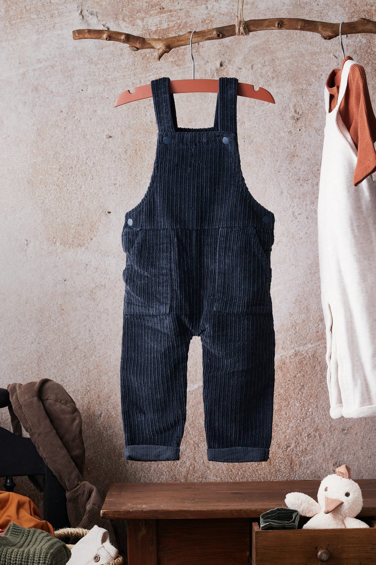Navy cord dungarees hanging on a wall with a teddy and wooden drawers below